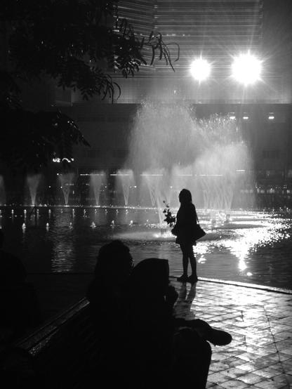 A person stands alone, holding a rose, in front of a pool of water jets in an urban square. The image is taken against the light in front of large spotlights, so we can only see er silhouette. (Version françæ) Æn personne pose, seulx, æn rose al main, devant æn bassin de jets d’eau sur æn place urbæn. On ne voit que saon silhouette car l’image est prix à contre-jour devant de granz projecteurs.