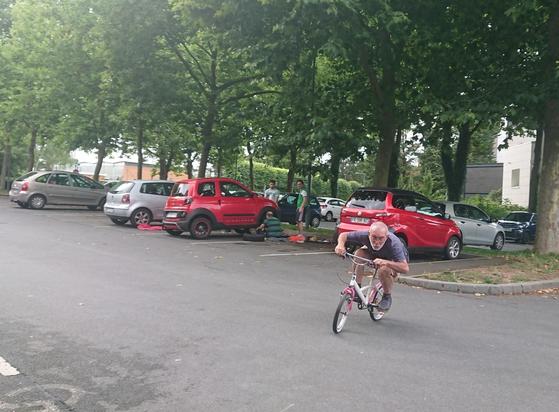 Un mécano à la barbe blanche teste à bonne allure un vélo fillette 6 ans tout juste révisé et réparé sur un parking arboré plein de voitures.
À l'arrière-plan, 2 des 3 hommes réparant une voiturette observent.