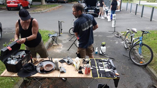 2 hommes rangent un établi plein d'outils, coupelles aimantés, café et cacahuettes sur LA place de ce grand parking où les véhicules à 4 roues n'ont pas accès. Où sont parqués 2 vélos néttoyés, des pompes et du matériel de nettoyage.