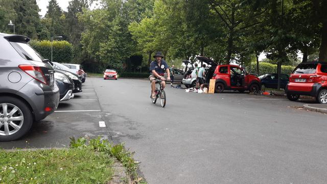 Un mécano barbu teste un vélo fillette 8 ans tout juste révisé et réparé sur un parking arboré rempli de voitures.