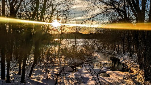 A soft blue late afternoon sky with streaky, gauzy thin clouds. We are standing along the bank of a small stream near its outlet into a small cove on a large lake. The stream and lake are surrounded by forests of leafless trees. An inch or so of wet snow coats the ground. The thin coating allows many of the ground's contours to show through. The stream and lake are mostly frozen over, with occasional dark patches of open water peeking through the snow covered ice. The gleaming pale orange sun is low above the horizon, giving the scene a soft golden glow mixed with deepening shadows. Tow bright horizontal rays extend left and right across the frame.