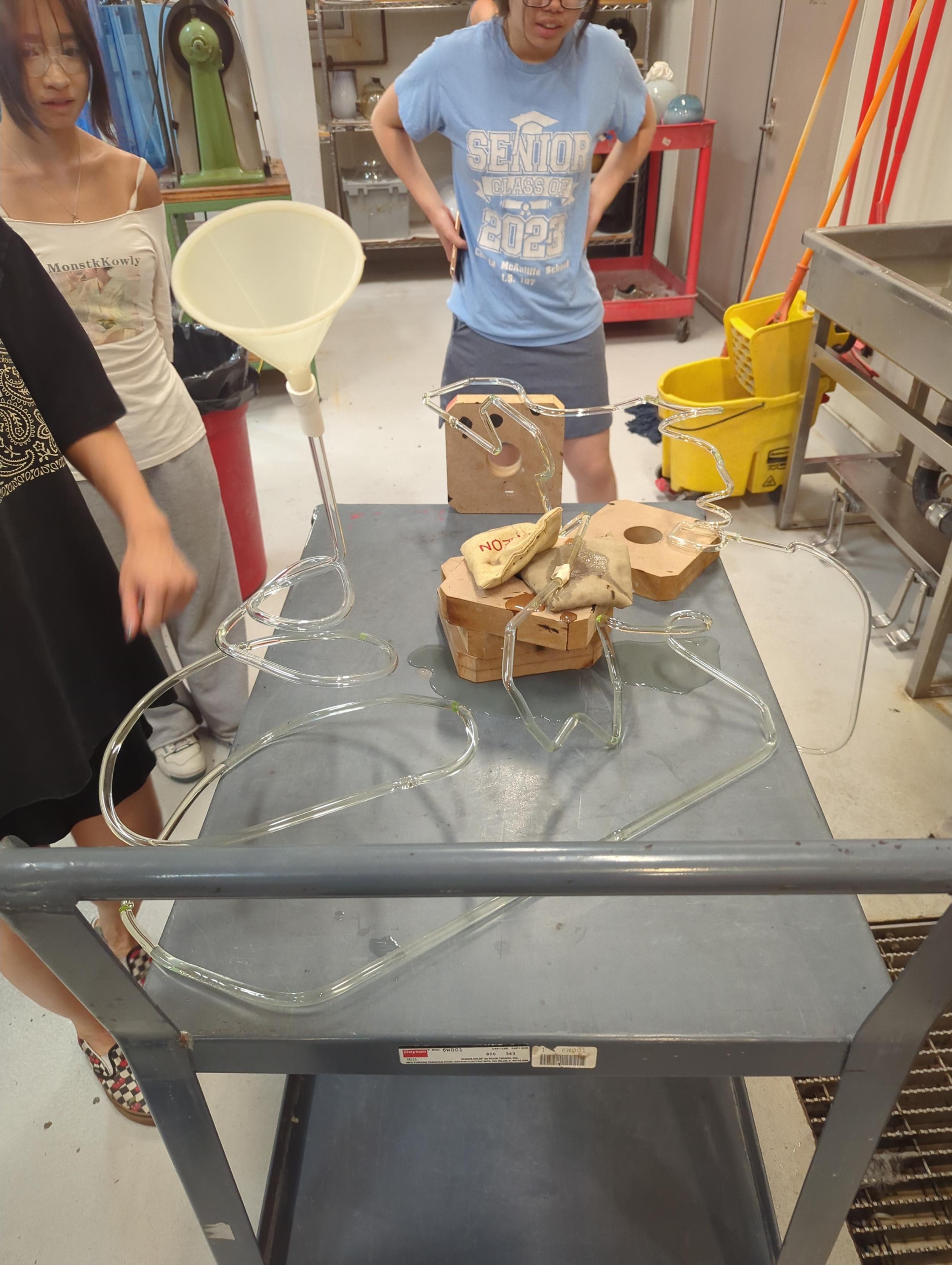 A glass roller coaster with bean bags strategically placed over the area of the rollercoaster with a crack in it. The glass tube rollercoaster is On a cart for mobility. Students wait to see if I will notice their cracked roller coaster