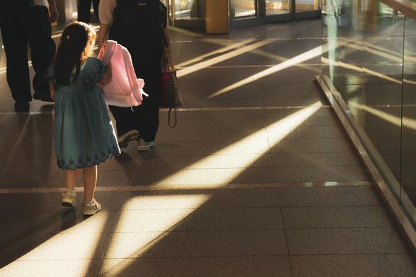 A young girl in a blue dress walks with an adult holding a pink item, possibly a bag, in a sunlit corridor. Strong diagonal sunlight creates