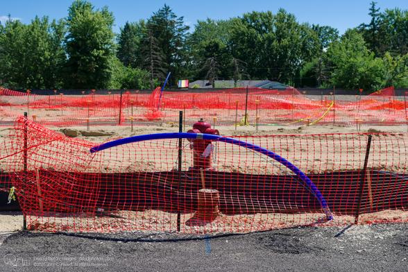 A red fire hydrant stands behind a section of orange construction fencing with a blue pipe running across it, curving up from the ground in a partial arch. There is fresh uneven asphalt in the foreground. The background shows a sandy construction site with more fencing, multiple capped rebar segments sticking up vertically, and trees.