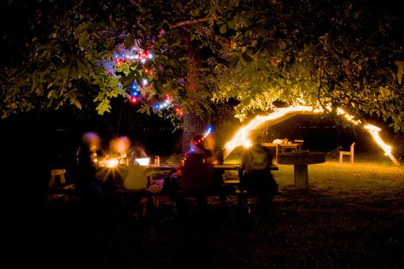 Ein Tisch an dem Leute sitzen die Verschwommen sind durch die Langzeitbelichtung. Dahinter ein Unterstand Beleuchtet und ein Baum mit Lichterkette. es ist dunkel.