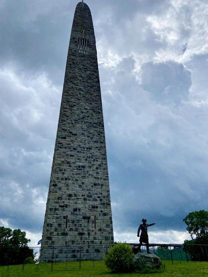 A 300-ft granite monument is silhouetted against a cloudy sky. A statue of a Minute Man points off to the right.