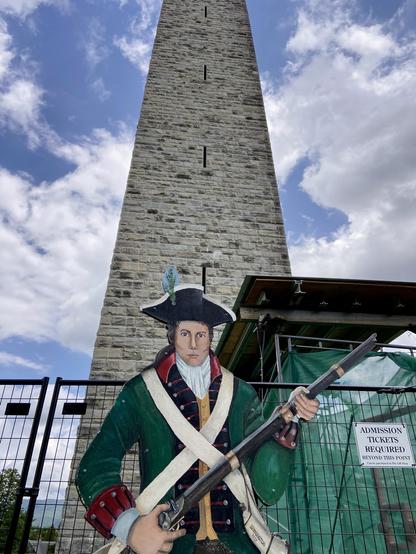 A wooden cutout of a Revolutionary War soldier waits for selfies at the base of the Bennington Monument. Some construction fence and green plastic surround the entrance.