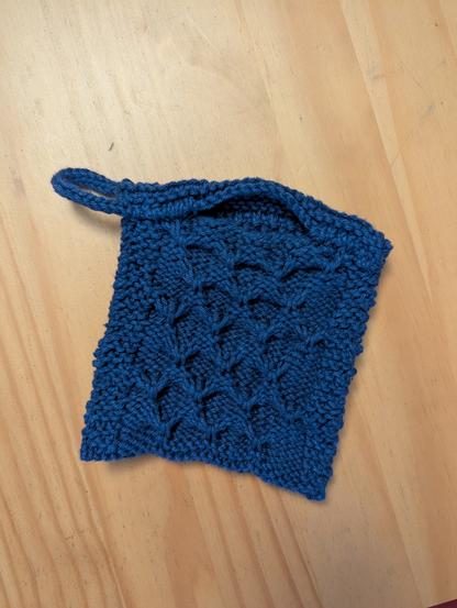 A navy blue knitted wash cloth sits on a wooden tabletop. It's hollow, and has a small folded over opening for holding a bar of soap. It has a textured trellis stitch surface perfect for scrubbing and creating bubbles. It also has an i-cord loop on one corner for hanging.
