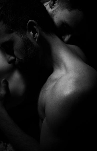 A black-and-white photograph of two shirtless men in an intimate pose, with one man's face resting near the other's neck and shoulder. The lighting emphasizes the contours of their bodies, creating a dramatic contrast of shadows and highlights. The image conveys vulnerability, connection, and raw emotion.