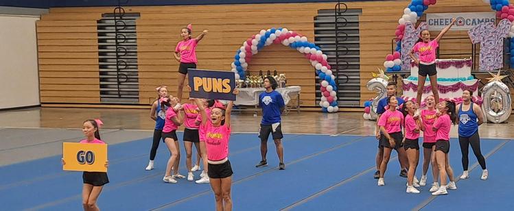 Middle school cheer time in pink shirts and black shorts. Finishing a routine the two girls in front are holding signs. The first says go. The second says puns