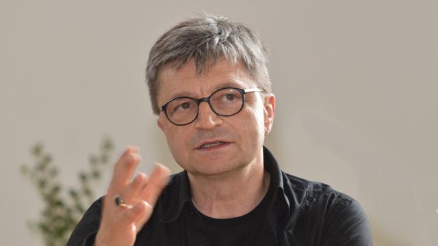 Porträt von Thomas Roscher mit kurzen, graumelierten Haaren und Brille. Er trägt eine schwarze, langärmelige Kleidung und eine runde Brille mit dunklem Gestell. Er hat einen ernsten Gesichtsausdruck und gestikuliert mit seiner rechten Hand, wobei ein Ring an seinem Ringfinger zu sehen ist. Der Hintergrund ist unscharf und hellbeige.