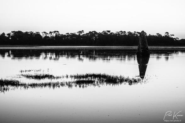 Photographie monochrome en noir et blanc, présentant un menhir cerné par la marée haute dans l'estuaire d'une rivière, à contre-jour, le soleil n'étant pas encore levé au-dessus des arbres de la rive opposée. 
La surface de l'eau est pratiquement lisse, ce qui permet le reflet du menhir, positionné sur le côté droit du cadrage à mi-hauteur, ainsi que des arbres de la rive opposée, en arrière-plan. 
Entre le photographe et le menhir, des pointes sommitales de plants de salicorne émergent encore de l'eau formant deux bandes, pratiquement horizontales, répondant en bas de la photo à la bande des arbres et de leur ombre de la rive opposée.
Aucun nuage dans le ciel, uniformément blanc ici