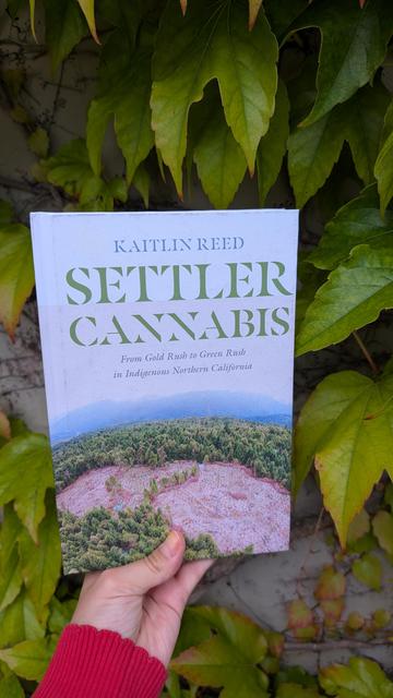 Holding the book "Settler Cannabis" by Kaitlin Reed in front of an overgrown wall