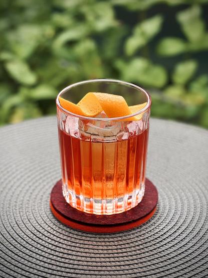 bright red cocktail with big rock of clear ice and orange twist garnish sitting on purple felt coaster with green herbs in the background