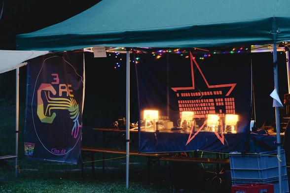 Die Tastaturstern und C3RE Banner hängen an der Seite eines Pavillon auf einer Wiese. Bei der Dämmerung kann scheinen durch den Tastaturstern-Banner die Tischlampen auf den Bierbänken.