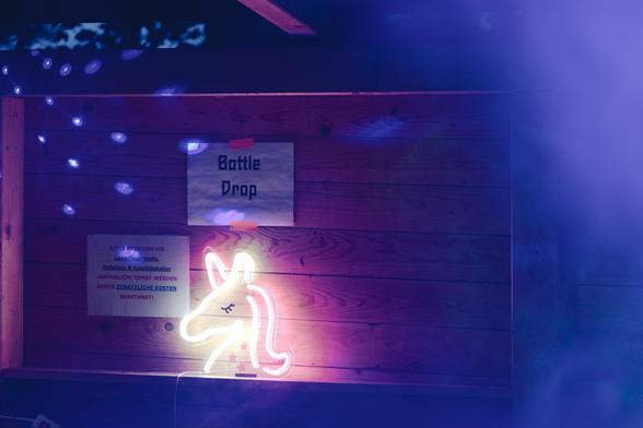 Die leuchtenden Umrisse eines Einhornkopf aus Leuchstoffröhren stehen in einem Holzverschlag unter einem "Bottle Drop" Schild. Der Rest des Bildes ist in Nebel und blauem Licht gehüllt.