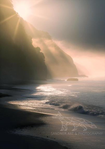 Golden sunrays pierce through a misty atmosphere, illuminating the coastline and ocean waves. The scene features a sandy beach meeting the gentle surf, with dark, rugged cliffs rising in the background. A bright highlight reflects on the water's surface from the sun's rays.
