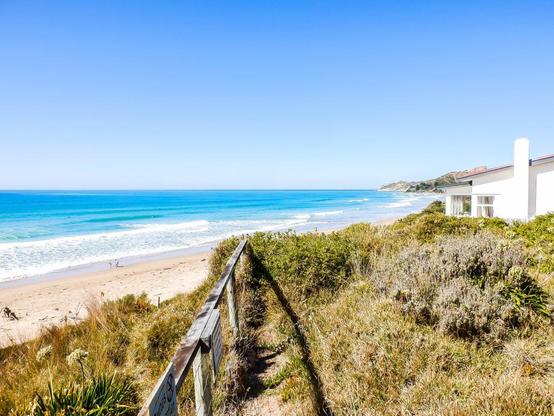 Beach view in Gisborner: sunny day, light blue ocean in the distance and white/yellow-ish sand on the beach. Dunes and track to the beach can be partially seen.