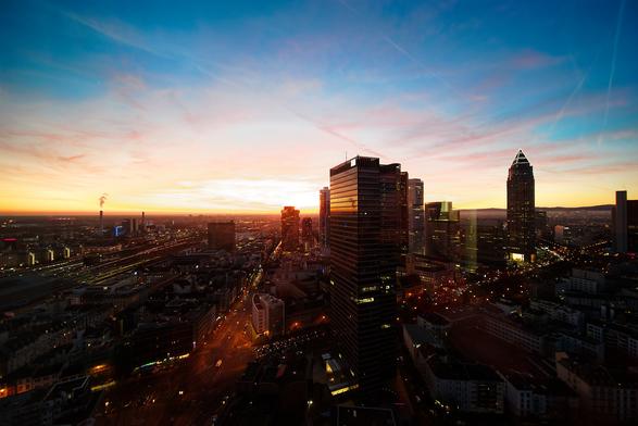 Blick von schräg oben auf das Frankfurter Bahnhofsviertel und den Messeturm. Oben der Abendhimmel (Blau, Rot und Gelbtöne). Unten die Gebäude und Straßenzüge (Schwarz und Orangetöne).