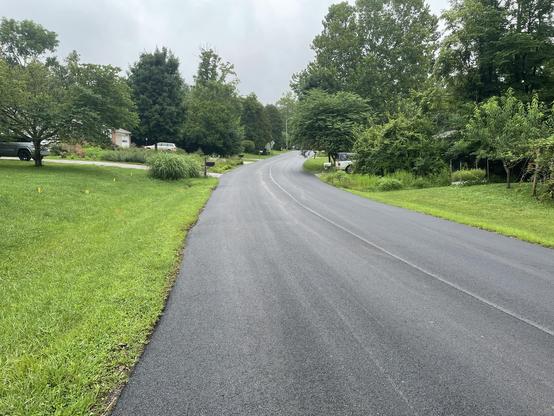 A freshly paved road in a quiet residential neighborhood. Mostly grass and trees visible beside the road; a couple houses peeling out from behind the trees.