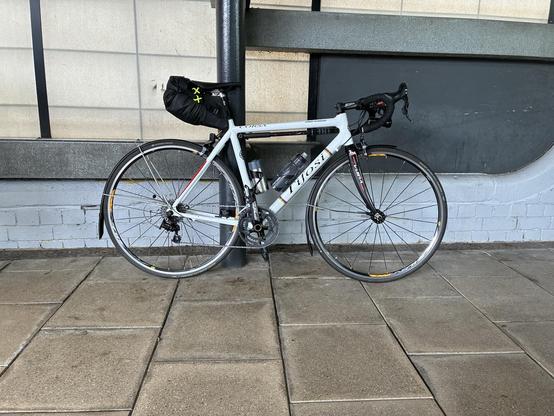 A white road bike is leaning against a pole. It has a black bag on the seat and a water bottle mounted on the frame, set on a tiled surface with a simple backdrop.