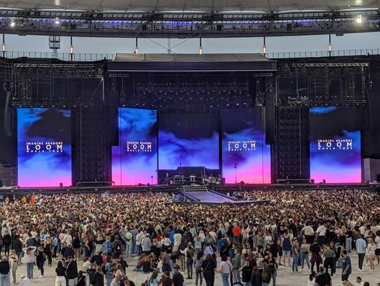 Große Konzertbühne mit leuchtenden Bildschirmen ‚Imagine Dragons Loom World Tour‘, Menschenmenge davor in Stadion.