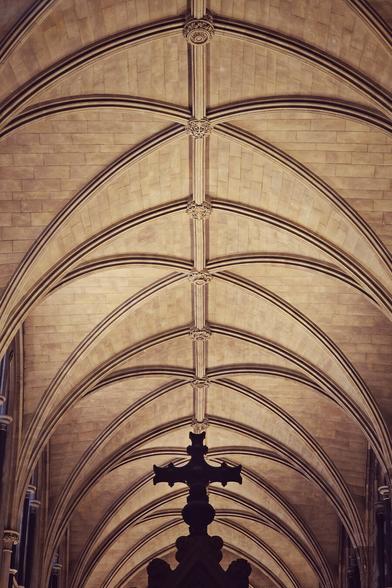 A low-angle, vertical shot of an ornate, vaulted stone ceiling within a grand, Gothic-style building. The ceiling features intricate rib vaulting with multiple arches converging, creating a sense of depth and height. The stone is a warm, light brown or beige colour, and there are decorative bosses at the intersections of the ribs. A dark, silhouetted cross or ornate finial hangs down from the centre of the frame, partially obscured by shadow. The lighting is somewhat dim, enhancing the historical and atmospheric feel of the interior.