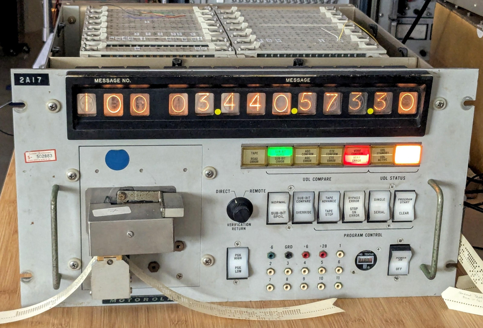 A gray box of electronics. It has thirteen large orange digits displayed at the top, with lights and switches underneath. These have labels such as Message, UDL Compare, AGC Confirm, and Program Start. A paper tape reader sticks out of the front, with a paper tape threaded through it. The unit is rack-mountable with two handles on the front.