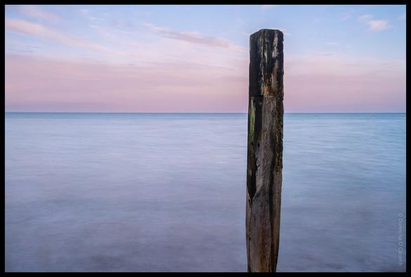 Long exposure photograph of a weathered wooden groyne post standing in calm water with soft pink and blue sunset colours reflected in the sea at Youghal beach, County Cork.