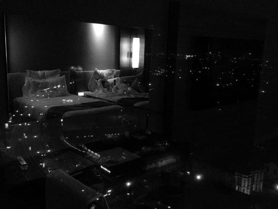 A hotel room's bay window reflects a person reading in bed against a backdrop of the city by night. (Version françæ) Reflet dans saon baie vitræ d’æn chambre d’hôtel avec æn personne lisant dans saon lit, sur fond de ville læ nuit.
