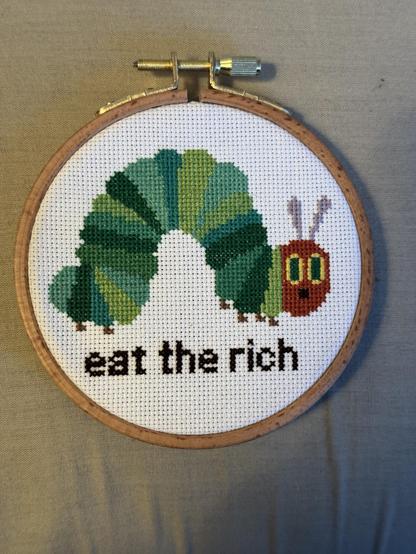 A small crossstitch hoop with a green caterpillar (that looks like Eric Carle's Very Hungry Caterpillar) with the words "eat the rich" underneath.