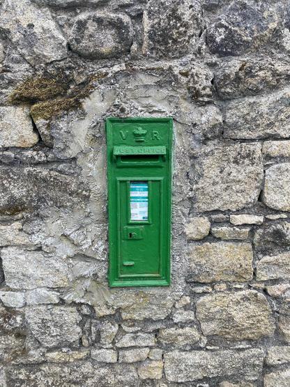 A small green cast iron post box is set in a stone wall. A glass window reveals the notice from An Post about the service and collection time in English and Irish. Above that, a slot for envelopes is sheltered by a flap with raised letters that say “post office”. Above that are a raised relief of a crown with the letters “V” and “R” on either side. Some moss grows on a few of the stones that are of different sizes but fit snugly together.
