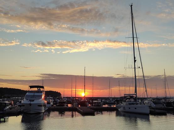 Una puesta de sol colorida sobre un puerto deportivo lleno de yates y veleros. El sol se encuentra en el horizonte, pintando el cielo con tonos anaranjados y rosados. Los barcos están amarrados a los muelles, con un gran velero en primer plano a la derecha. La escena es tranquila y pacífica.