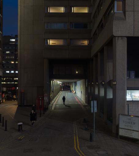 Evening view of concrete hotel, with a brightly lit road tunnel through it and a silhouetted figure in a coat standing at its entrance. There's a sign saying "The Tower Hotel"