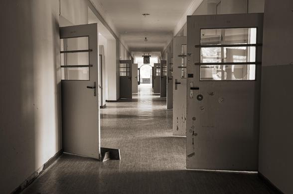 Dimly lit, monochromatic hallway lined with open doors on both sides. Each door has glass sections in the upper half, revealing bars. The floor is worn, and the corridor leads to a distant bright window, casting light along the path. Ceiling lights are evenly spaced, adding to the sparse illumination.
###
Schwach beleuchteter, einfarbiger Flur mit offenen Türen auf beiden Seiten. Jede Tür hat in der oberen Hälfte Glasabschnitte, die Gitterstäbe freilegen. Der Fußboden ist abgenutzt, und der Gang führt zu einem fernen, hellen Fenster, das Licht auf den Weg wirft. Die Deckenleuchten sind gleichmäßig verteilt, was die spärliche Beleuchtung noch verstärkt.