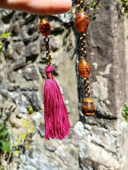 A set of worry beads on a long antiqued style chain, on the left of the image is a red silk tassel, the bead chain goes up and you can see several large oval glinting amber glass beads and vanishes into a hand before coming back down on the right showing a tigers eye pendant being used as an end.