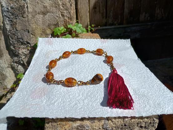 A chaplet style chain, there is a counter-clockwise coil starting on the right of the image with a red silk tassel and the body of chain and large amber beads going around in a spiral to a tigers-eye pendant at the centre.  It rests on a piece of white kitchen roll outside on a brick wall