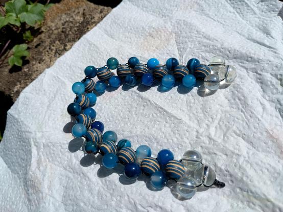 A Russian spine chetki style of beaded fidget, formed of a line of bigger 10mm beads down the centre, with smaller 8mm beads at their diagonal points.  The beading cord is thin black nanocord and is visible weaving down the sides and across the centres.  The ends are clusters of 10mm glass beads coloured to glint rainbow in the light.  It is resting on a piece of white kitchen roll coiled like a letter C.