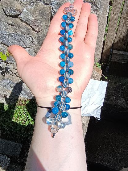 A Russian spine chetki style of beaded fidget, formed of a line of bigger 10mm beads down the centre, with smaller 8mm beads at their diagonal points.  The beading cord is thin black nanocord and is visible weaving down the sides and across the centres.  The ends are clusters of 10mm glass beads coloured to glint rainbow in the light. It is resting along the arm of a woman with pale pink skin, from index finger down to around the pulse point on the wrist where a tattoo is visible along with a thin black wrist bracelet.