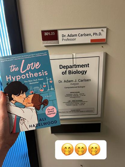 A hand holds up a copy of "The Love Hypothesis" by Ali Hazelwood in front of the door to Dr. Adam Carlsen's office on the set of the film adaptation. The door has a sign that says "Department of Biology. Dr. Adam J. Carlsen - Professor - Computational Biologist" with office hours listed.
