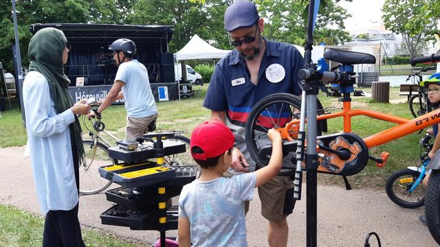 Un adulte aide un enfant, accompagné de sa mère,  à réparer son vélo fixé sur un pied d'atelier. à côté d'une servante à outil.