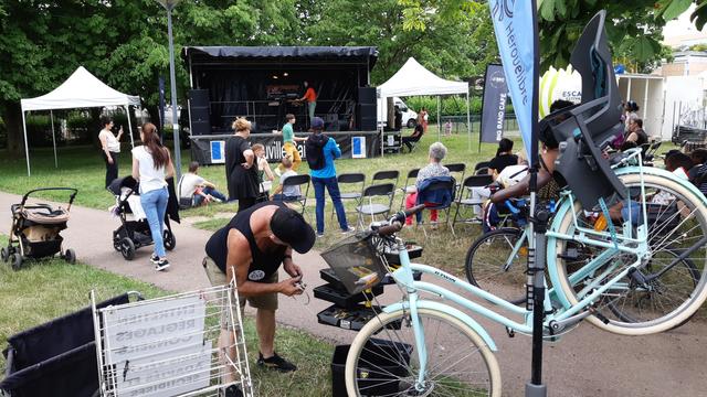 Un homme en pleine activité semble chercher l'outil adéquat dans une servante. Un vélo de femme blanc est fixé sur un pied d'atelier au premier plan avec l'oriflamme de l'association. À l'arrière les spectateurs commencent à prendre place face à la scène pour le concert en préparation.
