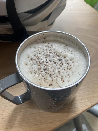 A reusable metal mug with steamed milk, and Hojicha tea and a sprinkling of freshly ground cardamom on top. A bicycle helmet is in the background. Both items are on a wooden table.