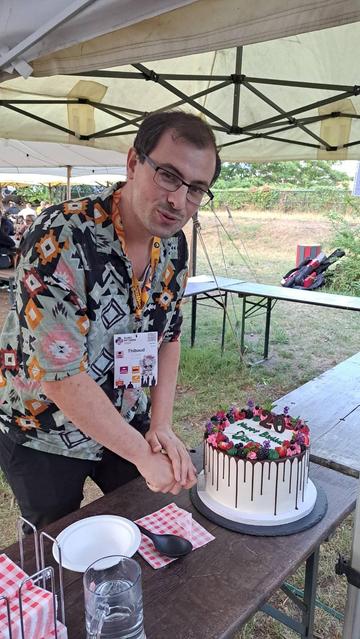 Django president Thibaud smiling, cutting a large cake that says '20 happy birthday django', outside under a tent