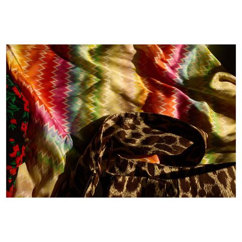 Flat closeup of various pieces of clothing in warm sidelight. Most of the frame is filled by a multi-coloured dress. At the bottom, a leopard print belt is resting on top of the dress. On the left edge, a dark dress with a red floral pattern is visible.