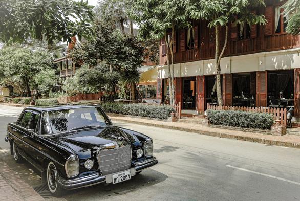 Photo shows a street with a black Mercedes-Benz vintage luxury car. The car has metal details and white rim tyres. In the background is a several two and three floor houses made in wood. The Architecture is a combination of Southeast Asian and French colonial building style. The houses are framed by and have trees, hedge rows and bushes making it into a green spot, nice town street. The car is from the 1960s, and the buildings may be from the same area. The reflections in the car’s metallic black paint tells of a sunny day with lightly clouded sky.