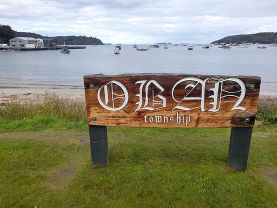 Le panneau du village d'Oban sur l'île Stewart, en Nouvelle-Zélande. Au fond se dessine l'océan, avec quelques bateaux dans le port.