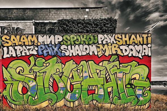 A vibrant graffiti mural dominates the foreground, featuring the word "PEACE" written in multiple languages across the top in yellow, green, and blue bubble letters. Below this, a large, intricate green graffiti tag with blue and pink accents covers the majority of the red brick wall. The wall stands on a grassy patch, with the weathered, grey building behind it contrasting with the colourful art. The sky above is cloudy and dramatic, adding to the urban feel of the scene.