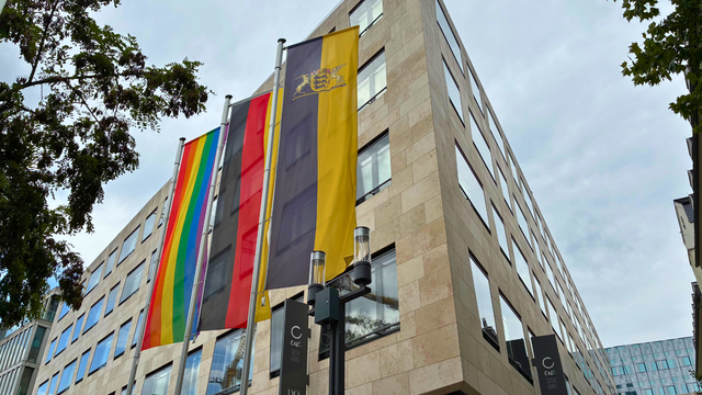 Foto von einem großen Gebäude. Davor stehen drei Flaggen: die Flagge von Baden-Württemberg, die Deutschland-Flagge und eine Regenbogenfahne.