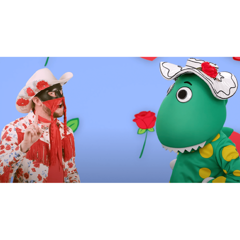 Orville Peck, wearing a nudie suit with red roses on a white background and a matching cowboy hat, glances to the camera as he wags a finger. He is dancing opposite someone in a green and yellow dinosaur suit. The background is lavender with cartoon red flowers.